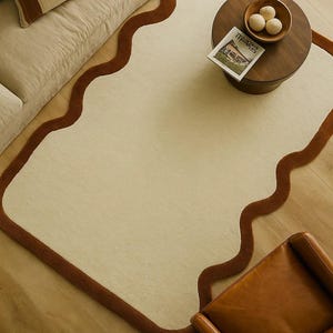 May include: A cream-colored rug with a wavy, brown border. The rug is placed on a wooden floor, with a sofa, coffee table, and leather chair visible. The coffee table has a wooden bowl and a magazine.