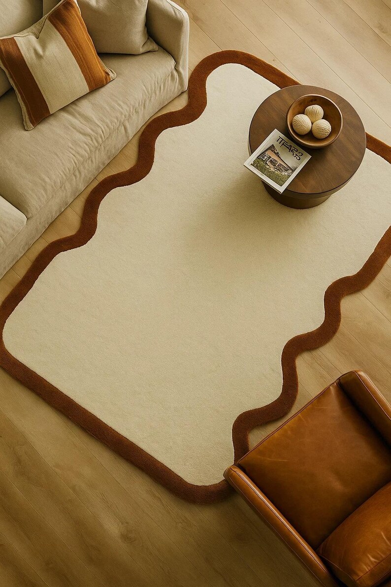 May include: A cream-colored rug with a wavy, brown border. The rug is placed on a wooden floor, with a sofa, coffee table, and leather chair visible. The coffee table has a wooden bowl and a magazine.