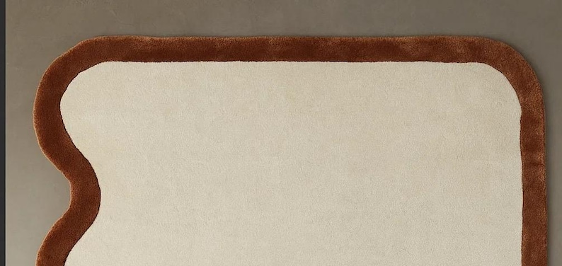 May include: A cream-colored rug with a brown border, shaped like a slice of bread. The rug has a soft, plush texture and a unique, playful design. This decorative rug is suitable for a bedroom or living room.