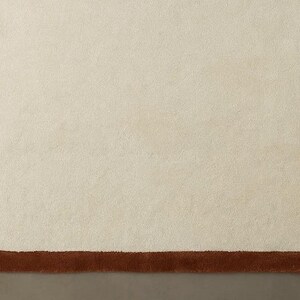 May include: A cream-colored rug with a unique, organic shape and a dark brown border. The rug's design features a soft, textured surface, creating a cozy and inviting aesthetic. This rug is suitable for living rooms and bedrooms.