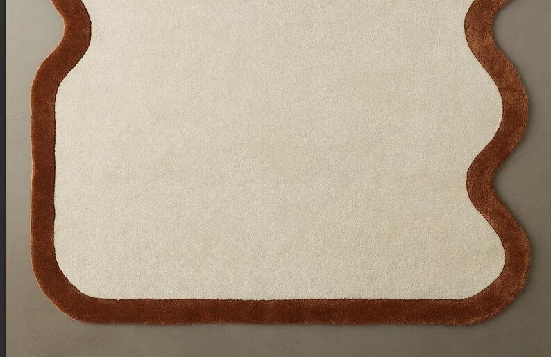 May include: A cream-colored rug with a unique, organic shape and a dark brown border. The rug's design features a soft, textured surface, creating a cozy and inviting aesthetic. This rug is suitable for living rooms and bedrooms.