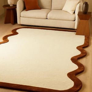 May include: Cream-colored area rug with a unique, wavy, brown border. The rug is placed in front of a beige sofa with brown and cream-colored pillows. A wooden side table and lamp are visible.