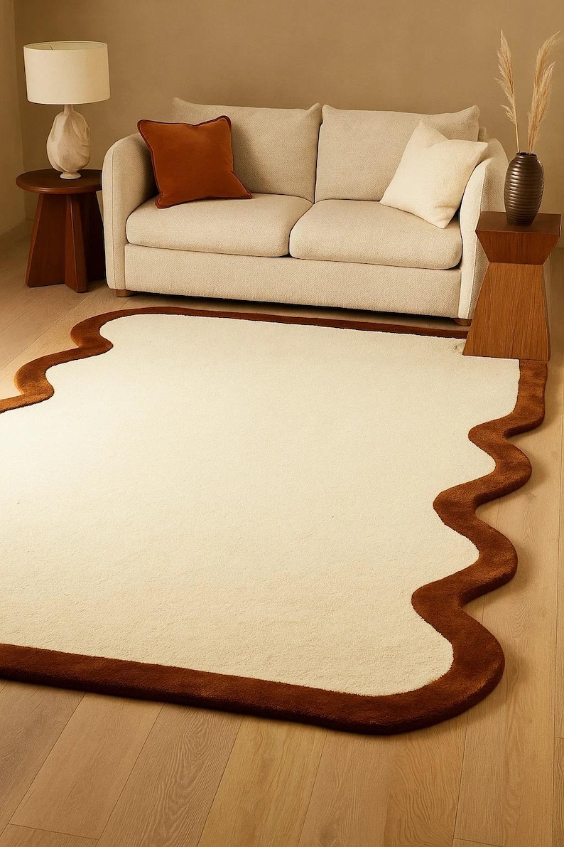 May include: Cream-colored area rug with a unique, wavy, brown border. The rug is placed in front of a beige sofa with brown and cream-colored pillows. A wooden side table and lamp are visible.