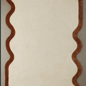 May include: A rectangular rug with a cream-colored center and a wavy, brown border. The rug has a soft texture and the text "Nazma arts" is visible in the upper right corner. The rug is designed for home decor.