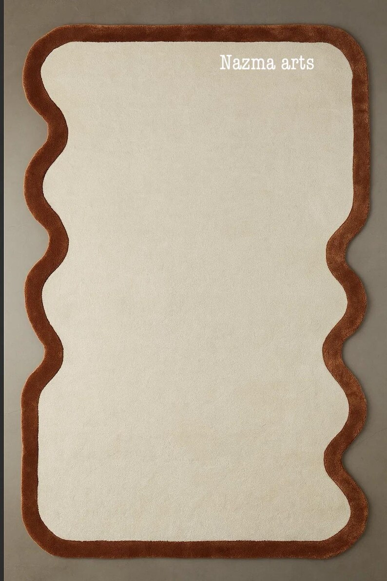 May include: A rectangular rug with a cream-colored center and a wavy, brown border. The rug has a soft texture and the text "Nazma arts" is visible in the upper right corner. The rug is designed for home decor.