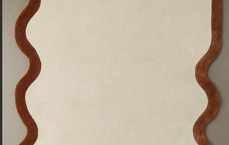 May include: A cream-colored rug with a wavy, brown border. The rug has a soft, textured appearance and a unique, organic shape. This decorative rug is suitable for a living room or bedroom.