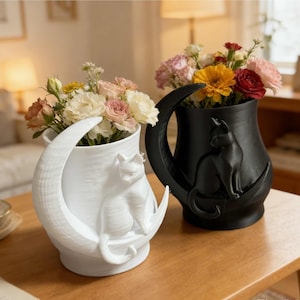 Op de afbeelding: Twee decoratieve vazen, een witte en een zwarte, elk met een kat op een halve maan. De witte vaas bevat lichtgekleurde bloemen, terwijl de zwarte vaas kleurrijke bloemen bevat. Beide vazen staan op een houten oppervlak.