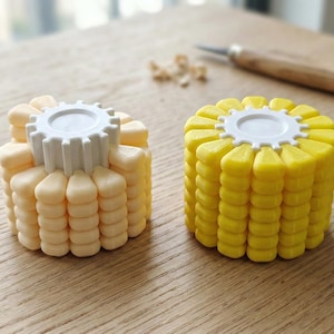 Puede incluir: Dos artilugios de cocina con forma de maíz. Uno es de color amarillo pálido, el otro es amarillo brillante. Ambos tienen una parte superior blanca en forma de engranaje. Una herramienta para tallar y virutas de madera están en el fondo.