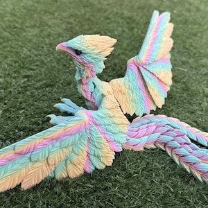 May include: A colorful fantasy bird figurine with outstretched wings and a long, flowing tail. The bird features pastel shades of yellow, pink, and blue. The figurine is set against a green, grassy background.