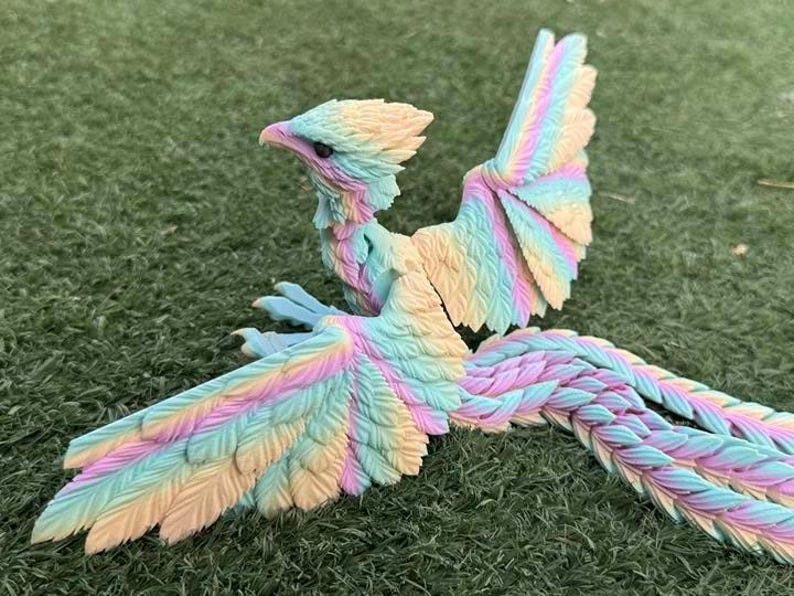 May include: A colorful fantasy bird figurine with outstretched wings and a long, flowing tail. The bird features pastel shades of yellow, pink, and blue. The figurine is set against a green, grassy background.