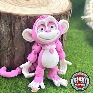 May include: A pink and white articulated monkey figurine with a smiling face, large ears, and a curled tail. The monkey is posed on a bed of green grass and in front of a tree trunk. The Flexi Factory logo is visible.