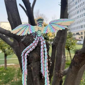 May include: A colorful, handcrafted bird sculpture with outstretched wings and flowing tail feathers. The sculpture features pastel hues of pink, blue, and yellow, creating a whimsical and artistic design. It is perched on a tree branch, showcasing its intricate details.