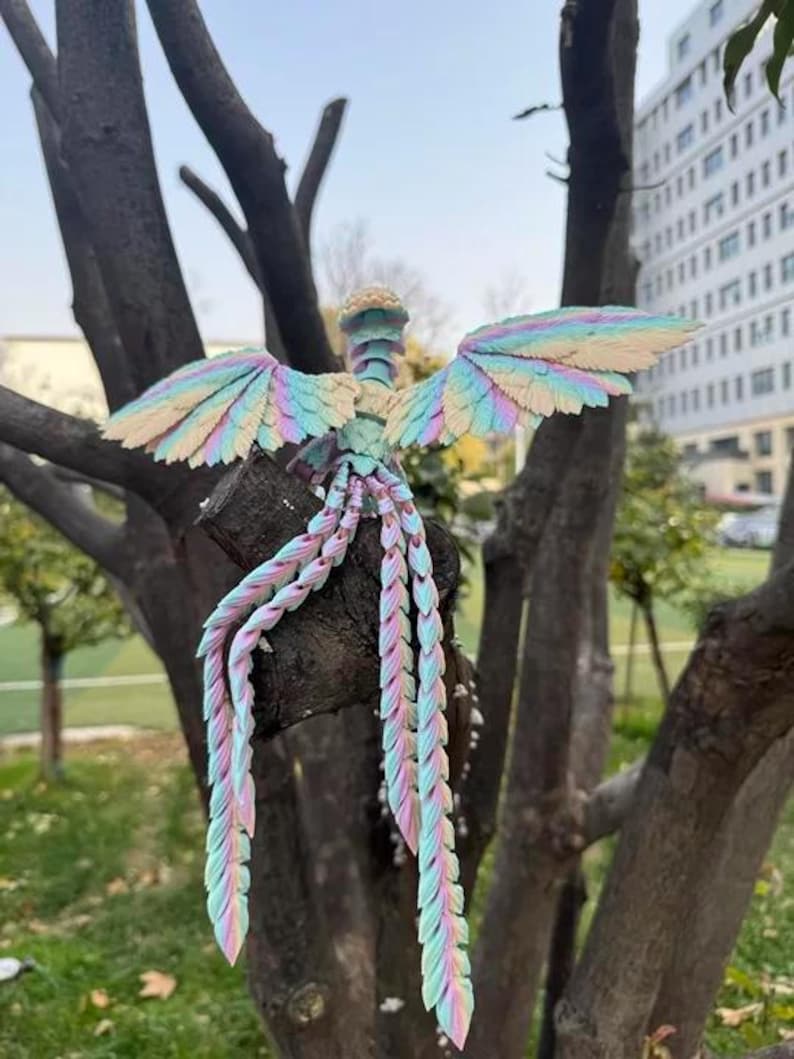 May include: A colorful, handcrafted bird sculpture with outstretched wings and flowing tail feathers. The sculpture features pastel hues of pink, blue, and yellow, creating a whimsical and artistic design. It is perched on a tree branch, showcasing its intricate details.
