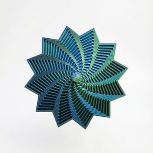 Puede incluir: Una obra de arte en capas con forma de estrella y diseño en espiral. La obra presenta tonos de azul y verde, creando un efecto degradado. La pieza está sobre un fondo blanco, resaltando su intrincada forma geométrica.