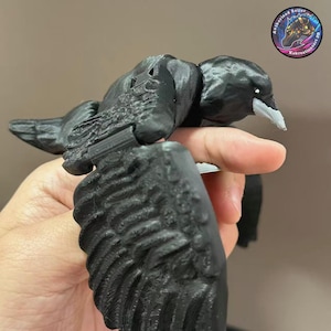 Puede incluir: Un pájaro negro impreso en 3D con alas extendidas y un pico blanco. Las alas del pájaro están detalladas con plumas texturizadas. El pájaro se sostiene en una mano, mostrando su tamaño y diseño. El pájaro tiene un pequeño ojo blanco.