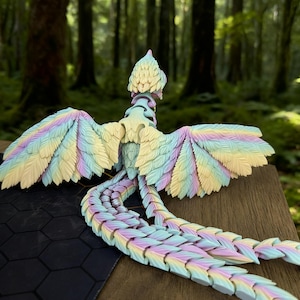 May include: A pastel-colored articulated dragon figurine with a detailed feather design. The dragon features a rainbow of colors, including yellow, blue, and purple. The figurine is posed with its wings spread and tail extended, set against a blurred forest background.
