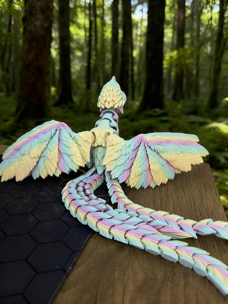 May include: A pastel-colored articulated dragon figurine with a detailed feather design. The dragon features a rainbow of colors, including yellow, blue, and purple. The figurine is posed with its wings spread and tail extended, set against a blurred forest background.