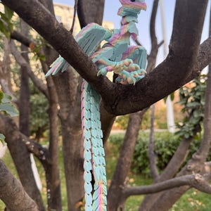 May include: A colorful, articulated dragon figurine perched on a tree branch. The dragon features a pastel color scheme with shades of blue, pink, yellow, and green. Its wings are spread, and its long tail hangs down. The dragon is a fantasy-themed item.