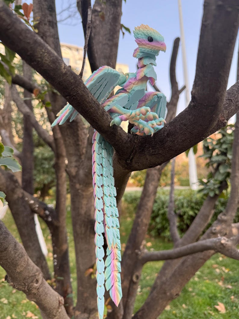 May include: A colorful, articulated dragon figurine perched on a tree branch. The dragon features a pastel color scheme with shades of blue, pink, yellow, and green. Its wings are spread, and its long tail hangs down. The dragon is a fantasy-themed item.
