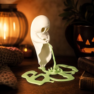 May include: A white ghost figurine with a smooth, rounded head and draped body, depicted in a standing position. The ghost is shown emitting a light green liquid stream that pools into a flat, irregular puddle shape on the surface below.