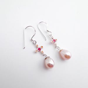 May include: A pair of silver drop earrings with pink, teardrop-shaped pearls. The earrings have a decorative cross-shaped design with red and orange accents. The hooks are silver.