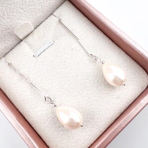 Puede incluir: Pendientes colgantes con perlas rosas claras en forma de pera suspendidas de finas cadenas de plata. Los pendientes se exhiben dentro de un joyero rosa con un interior blanco y afelpado, creando una presentación elegante.