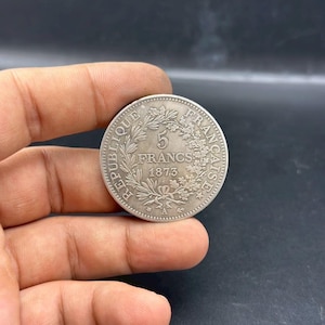 以下が含まれることがあります： 中央に「5 FRANCS 1873」の文字、周囲にリースと「REPUBLIQUE FRANCAISE」の文字が刻まれた銀貨。 暗い背景を背景に、2つの手でコインが持たれています。 コインは丸く、詳細なデザインが施されています。