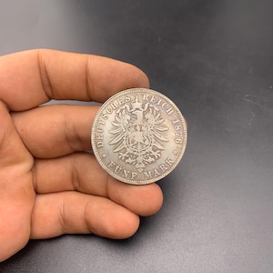 May include: A silver coin with intricate detailing, featuring an eagle emblem and the text "DEUTSCHES REICH 1876". The coin is held in a hand, showcasing its size and design. The coin reads "FÜNF MARK".