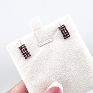 Puede incluir: Un par de pendientes de botón rectangulares. Cada pendiente presenta una rejilla de pequeñas piedras preciosas de color rojo oscuro engastadas en un metal plateado. Los pendientes se exhiben sobre una superficie blanca texturizada.