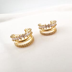 Może przedstawiać: Dwa złote nauszniki. Każdy nausznik ma trzy warstwy. Górna warstwa jest ozdobiona prostokątnymi, przezroczystymi kryształami. Środkowa warstwa jest gładka, złota, a dolna warstwa jest ozdobiona małymi, okrągłymi kryształami. Tło jest białe.