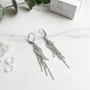 May include: Silver drop earrings with a wing and chain design. The earrings feature a dangling chain and wing-shaped details. A white gift box with a silver ribbon is in the background. The text reads "Sterling Silver Wing & Chain Drop Earrings - Elegant Design -"
