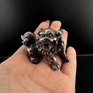 Peut inclure: Une petite figurine en métal argenté représentant une créature lion mythique, avec des détails sculptés complexes sur la crinière, la queue et le visage, une gueule ouverte montrant les dents et une finition texturée et patinée sur tout le corps.