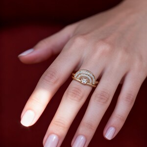 Op de afbeelding: Een gouden ringenset met een diamant en een reeks kleinere stenen gerangschikt in een boogontwerp. De ring zit aan een hand met verzorgde nagels, tegen een rode achtergrond.