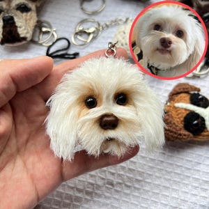 May include: A handmade white dog keychain with a fluffy texture, brown nose, and dark eyes. The keychain is attached to a silver metal ring. Other dog-themed keychains are visible in the background.