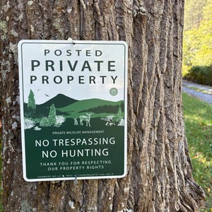May include: A rectangular white and dark green sign attached to a tree trunk. The text reads POSTED PRIVATE PROPERTY, PRIVATE WILDLIFE MANAGEMENT, NO TRESPASSING NO HUNTING, and THANK YOU FOR RESPECTING OUR PROPERTY RIGHTS with a deer graphic.