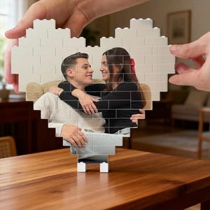 May include: A white heart-shaped photo frame made of interlocking blocks, with a couple embracing inside. The frame is on a wooden table, held by hands. The photo shows a man and woman smiling at each other.