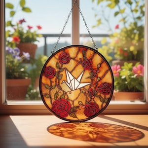 Op de afbeelding: Ronde glas-in-loodraamkunst met rode rozen en een witte origami-kraanvogel. Het raam wordt opgehangen door een ketting en heeft een warme, amberkleurige achtergrond. Het raam wordt tentoongesteld voor een raam met potplanten.