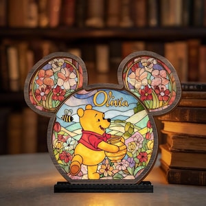 Op de afbeelding: Een Winnie de Poeh glas-in-lood lamp in de vorm van Mickey Mouse. De lamp heeft een kleurrijk bloemenontwerp en de naam "Olivia". Poeh draagt een honingpot. De lamp staat op een zwarte voet.