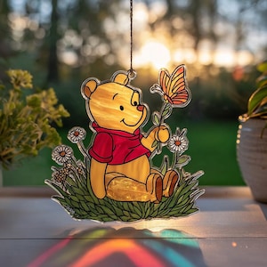 May include: A stained glass Winnie the Pooh suncatcher. The yellow bear is wearing a red shirt and holding a flower with a butterfly. The suncatcher is surrounded by green grass and white daisies. The background is a blurred view of a sunset.
