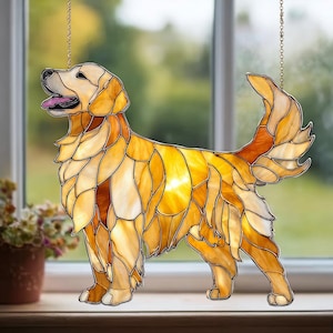 Peut inclure: Décoration de chien golden retriever en vitrail. Le chien est représenté debout, la langue sortie. Les pièces de verre sont dans des tons de jaune, d'orange et de marron, avec une chaîne dorée pour l'accrocher. Le chien est devant une fenêtre.