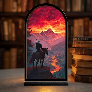 May include: A framed art piece depicting a figure on horseback overlooking a mountain landscape at sunset. The artwork features a vibrant color palette of red, orange, and purple, with a river winding through the valley. The frame is black.