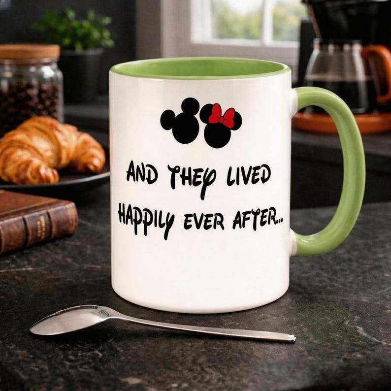 Puede incluir: Taza de cer&aacute;mica blanca con asa y borde interior verdes. La taza presenta siluetas negras de Mickey y Minnie Mouse, con Minnie luciendo un lazo rojo. El texto "AND THEY LIVED HAPPILY EVER AFTER..." est&aacute; impreso debajo.