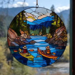 Pode incluir: Painel redondo de vitral retratando uma cena de rio com dois caiaques. A obra apresenta um rio azul, árvores verdes e montanhas em tons de azul, marrom e amarelo. Duas pessoas estão nos caiaques, usando chapéus e coletes salva-vidas.