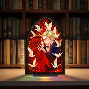 May include: A stained glass panel showing a couple in an embrace, with origami cranes around them. The artwork features a red-haired woman and a blonde man, set against a red background. The panel is framed in black and sits on a wooden base.