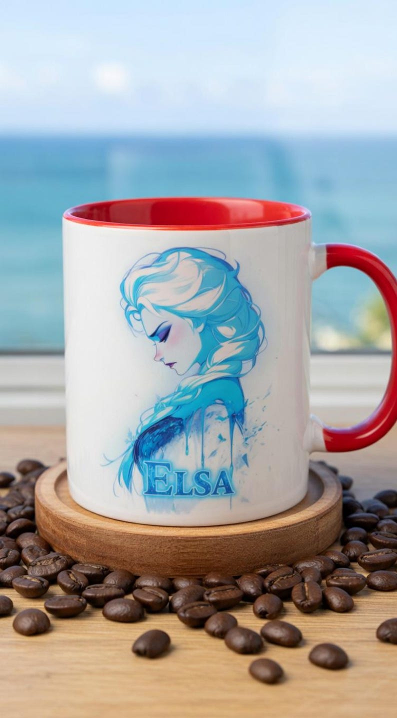 May include: White ceramic mug with a red interior and handle. The mug features a blue and white illustration of a woman with the name "ELSA" in blue. The mug is on a wooden coaster, surrounded by coffee beans.