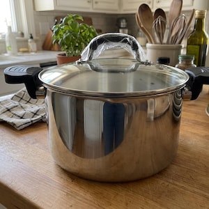 May include: A stainless steel cooking pot with a glass lid and black handles. The pot is sitting on a wooden surface. A stack of plates and a checkered towel are nearby. Kitchen utensils and a plant are in the background.