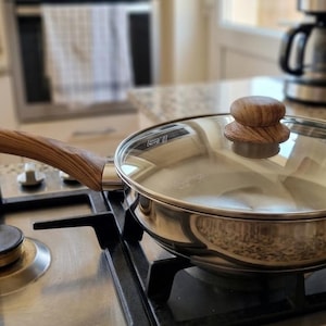 May include: Stainless steel frying pan with a glass lid and wooden handle, resting on a gas stove. The handle and lid knob are made of light brown wood. The pan is on a black stove top.