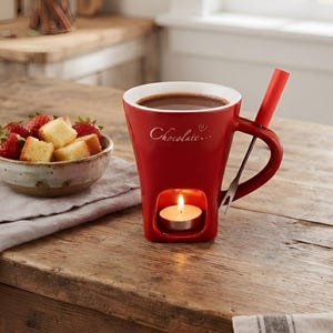May include: A red ceramic chocolate fondue mug with a white interior and the word Chocolate written in white script on the front. The mug features a hollow base containing a lit tea light candle and a red fondue fork resting in the handle.