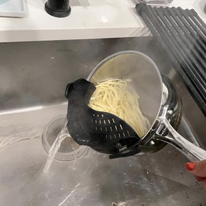 May include: A stainless steel saucepan containing cooked spaghetti noodles with a black silicone clip-on strainer attached to the rim, allowing hot water to drain out into a sink while keeping the pasta inside the pot during the process.