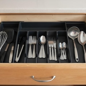 May include: A dark gray plastic kitchen drawer organizer tray with multiple compartments containing various stainless steel utensils including a whisk, spatula, tongs, forks, spoons, knives, a ladle, and a large serving spoon inside a wooden drawer.